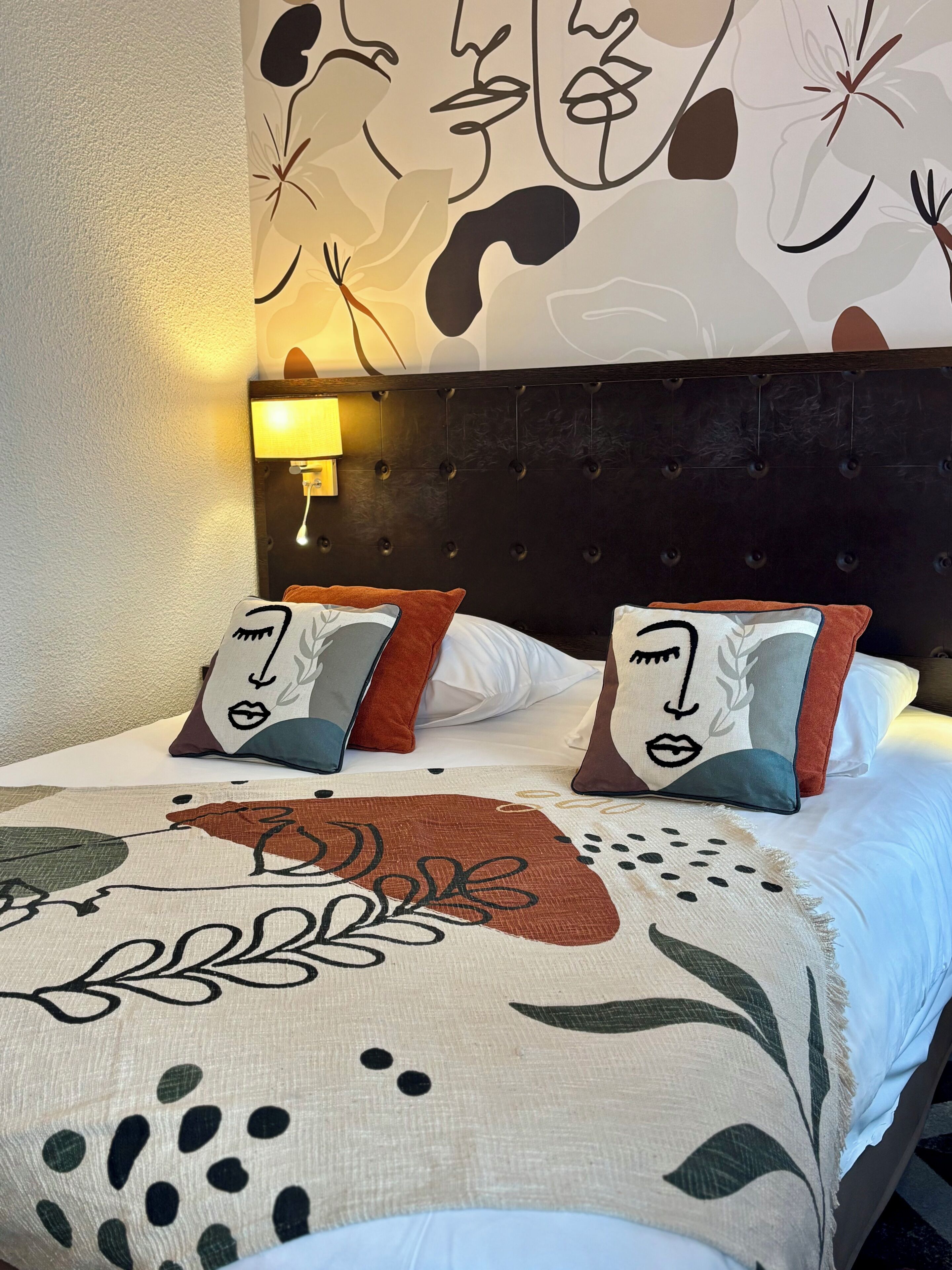 A bed with a patterned bedspread and two decorative pillows, a wall-mounted lamp, and a tufted headboard.