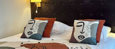 Una cama con una colcha estampada y dos almohadas decorativas, una lámpara de pared y un cabecero capitonado.