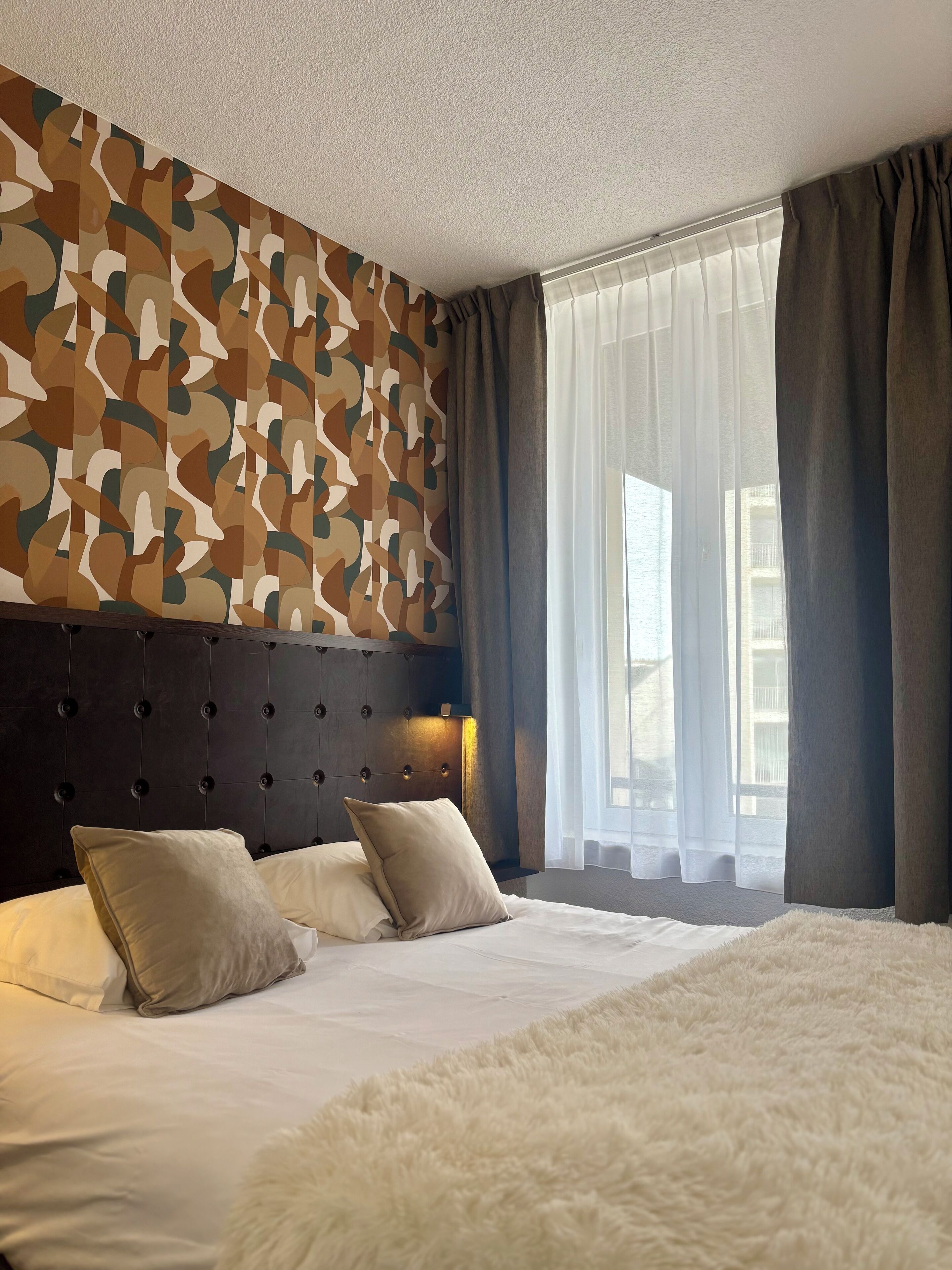 A hotel room with a tufted headboard, a large bed with white linens, beige pillows, and a fluffy white rug.