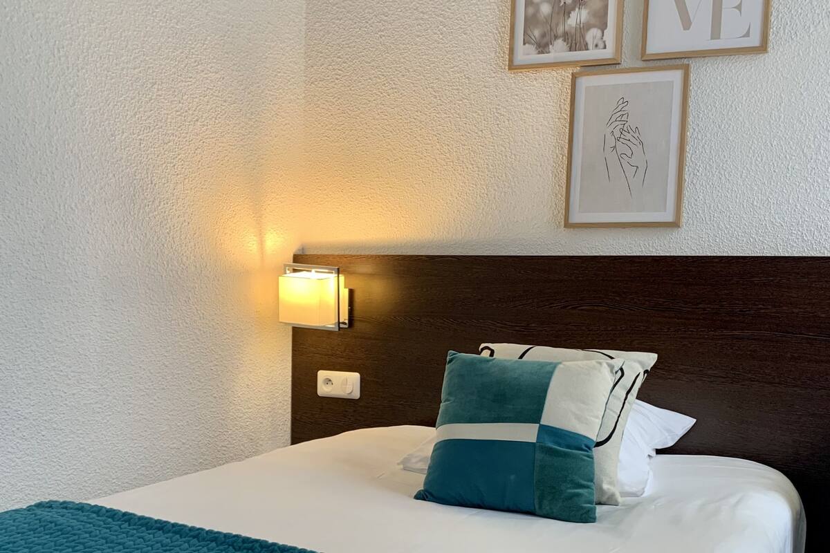 A neatly made bed with a teal blanket and pillows, a wall-mounted light, and framed artwork above it.