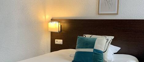 Una cama bien hecha con un edredón y almohadas color turquesa, una lámpara de pared y cuadros colgados arriba.