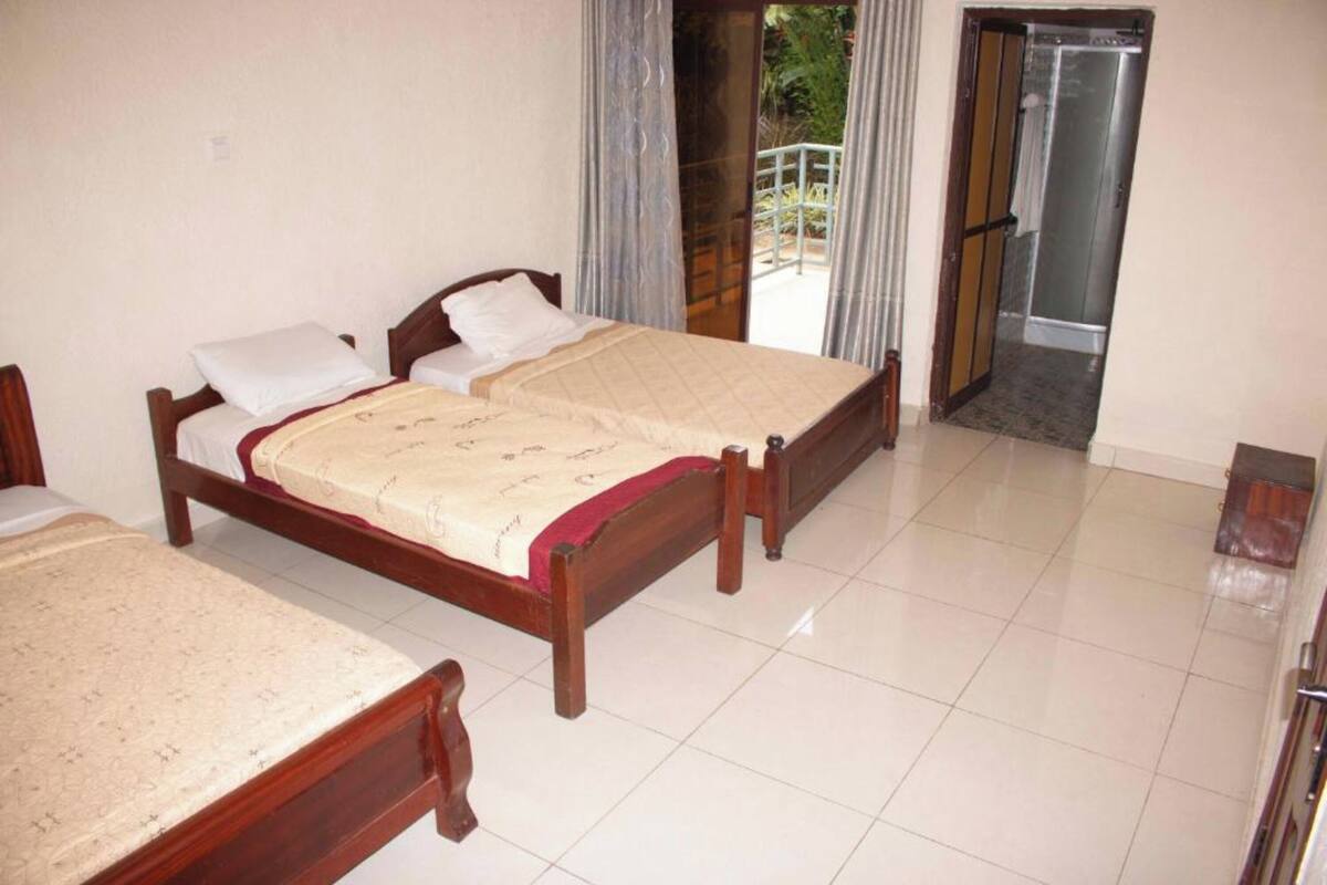 Habitación de hotel con dos camas de madera, un balcón con cortinas y una puerta corrediza de cristal que da a una zona de ducha.