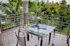 A glass table with a laptop and teapot on a balcony with a metal railing, surrounded by lush greenery.