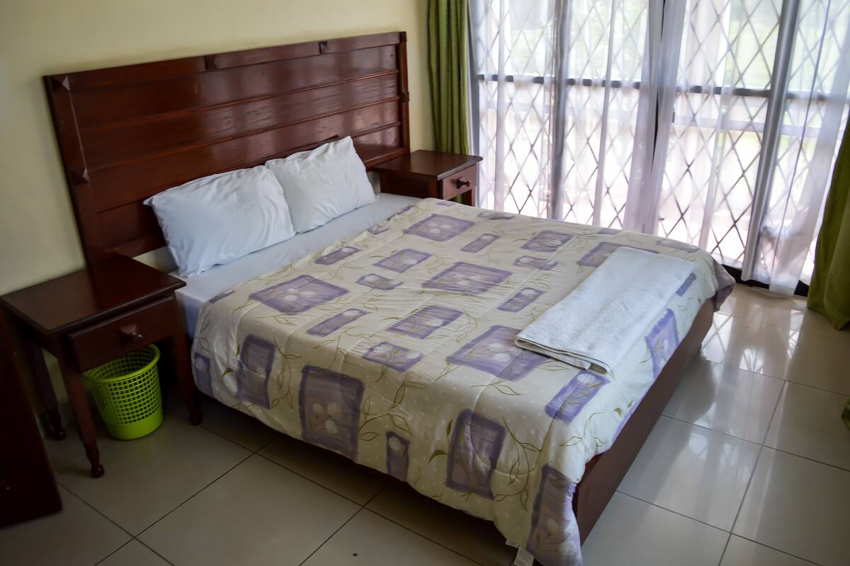 A neatly made bed with a patterned bedspread, a wooden headboard, two nightstands, a green basket, and a view through a window with curtains.