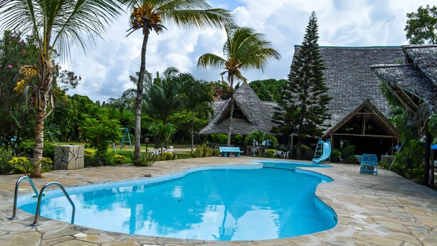 A swimming pool with stone tiles, surrounded by palm trees and a thatched-roof building. - Greenwood Resort (Mtwapa)
