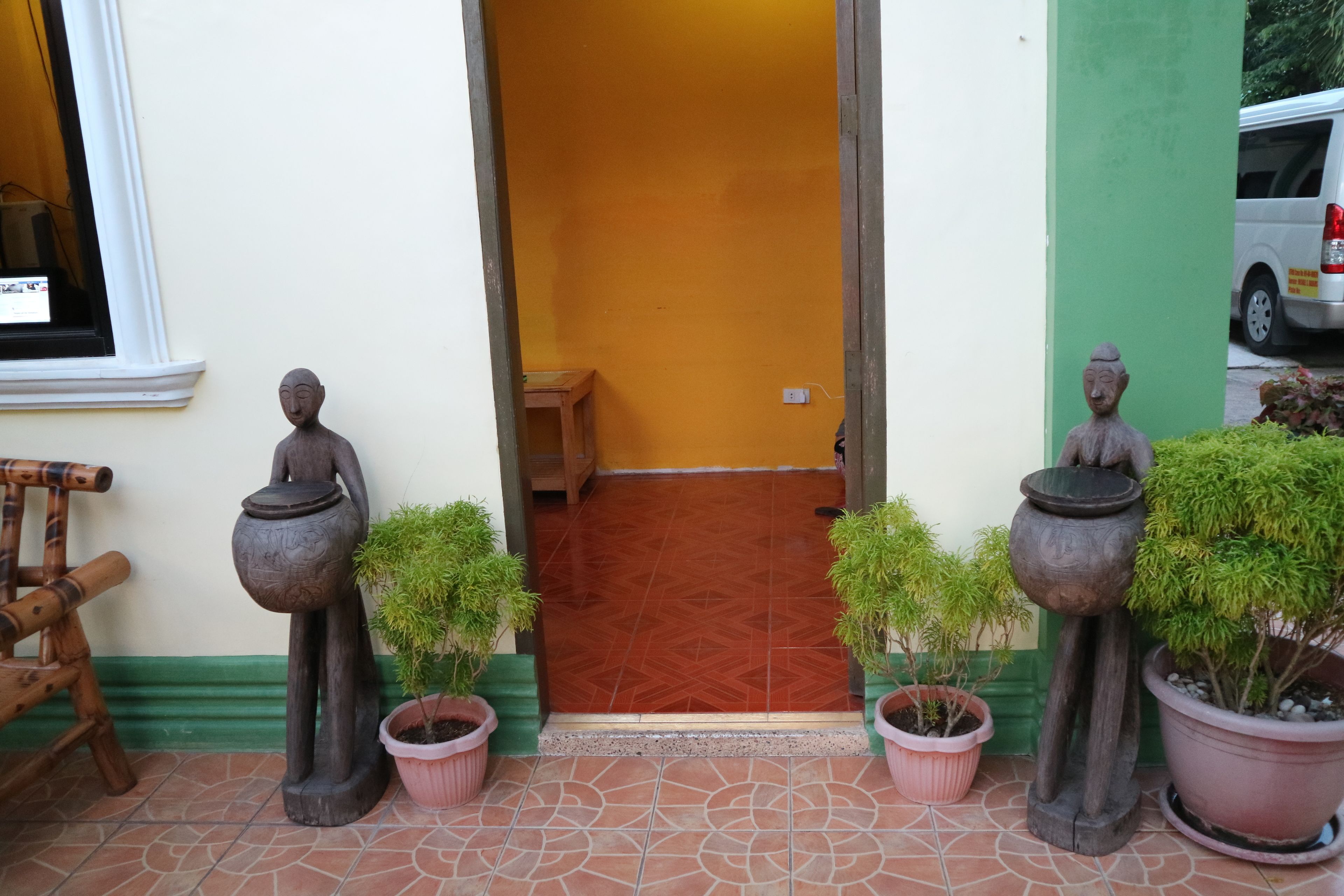 Un vano con dos estatuas sosteniendo macetas, flanqueado por plantas en macetas, que da paso a una habitación con suelo estampado y un banco.