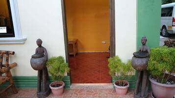 Un vano con dos estatuas sosteniendo macetas, flanqueado por plantas en macetas, que da paso a una habitación con suelo estampado y un banco.