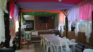 Un área de comedor al aire libre techada con una mesa larga y sillas, un televisor montado en la pared y un letrero que indica servicio propio.