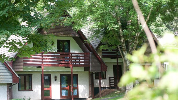 A two-story house with a balcony, surrounded by trees. - Tolroy Manor Holiday Park (Hayle)