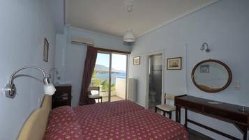 A bedroom with a bed, a desk, a chair, a mirror, and a view of the sea.