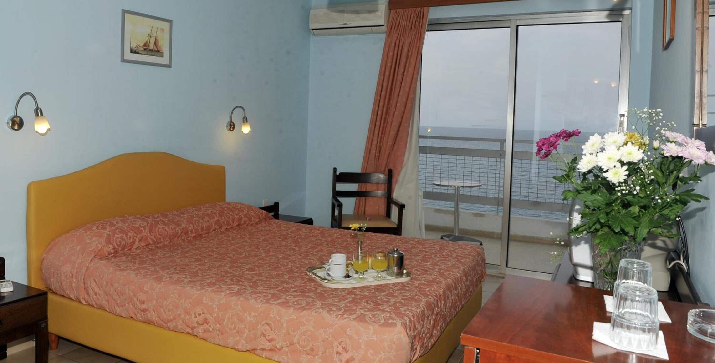 A hotel room with a bed, a small table with a tray of breakfast, a chair, a desk with a vase of flowers, and a view of the sea through a large window.