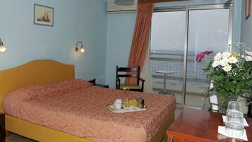 Quarto de hotel com cama, uma pequena mesa com bandeja de café da manhã, uma cadeira, escrivaninha com vaso de flores e vista para o mar através de uma grande janela.