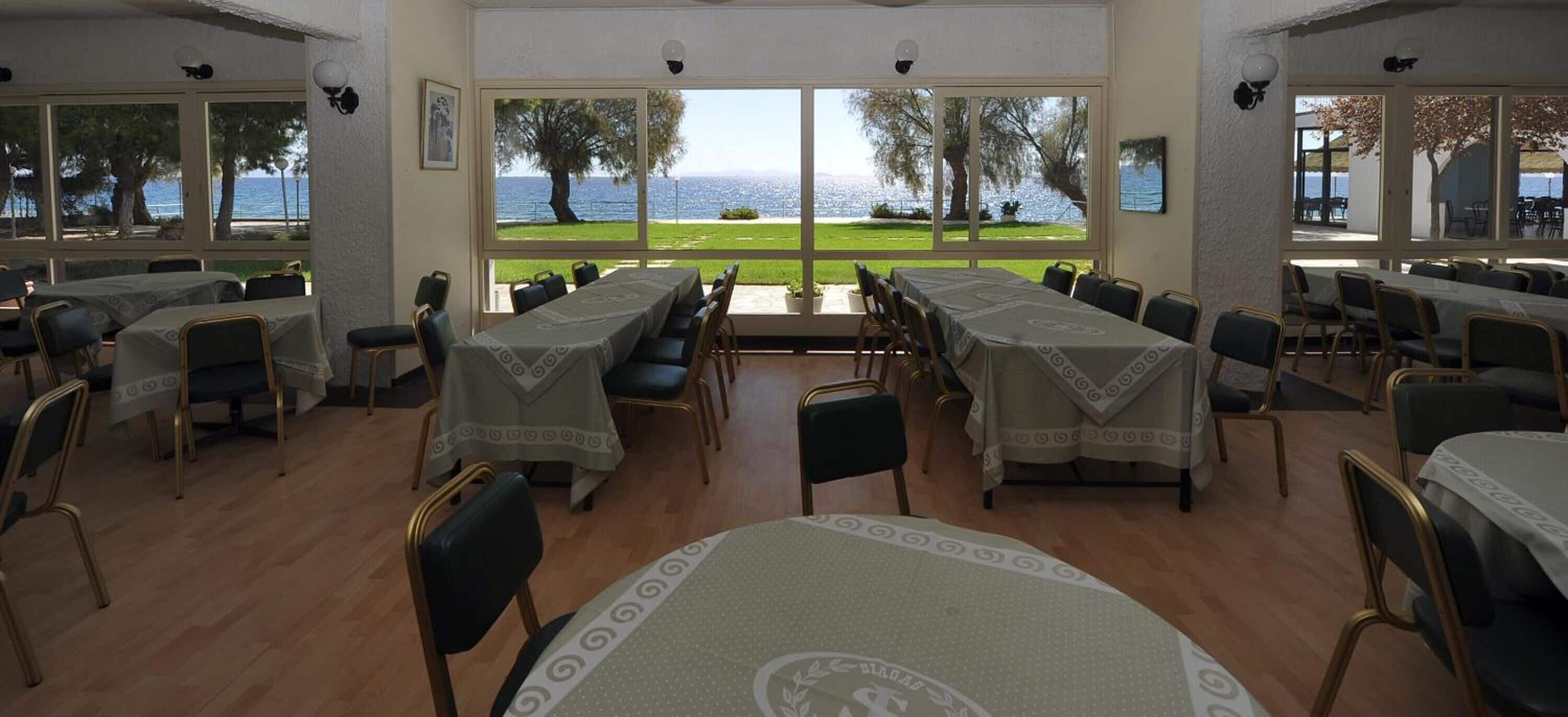 Un comedor con mesas y sillas, ventanas amplias con vistas al mar y árboles, y suelo de madera.