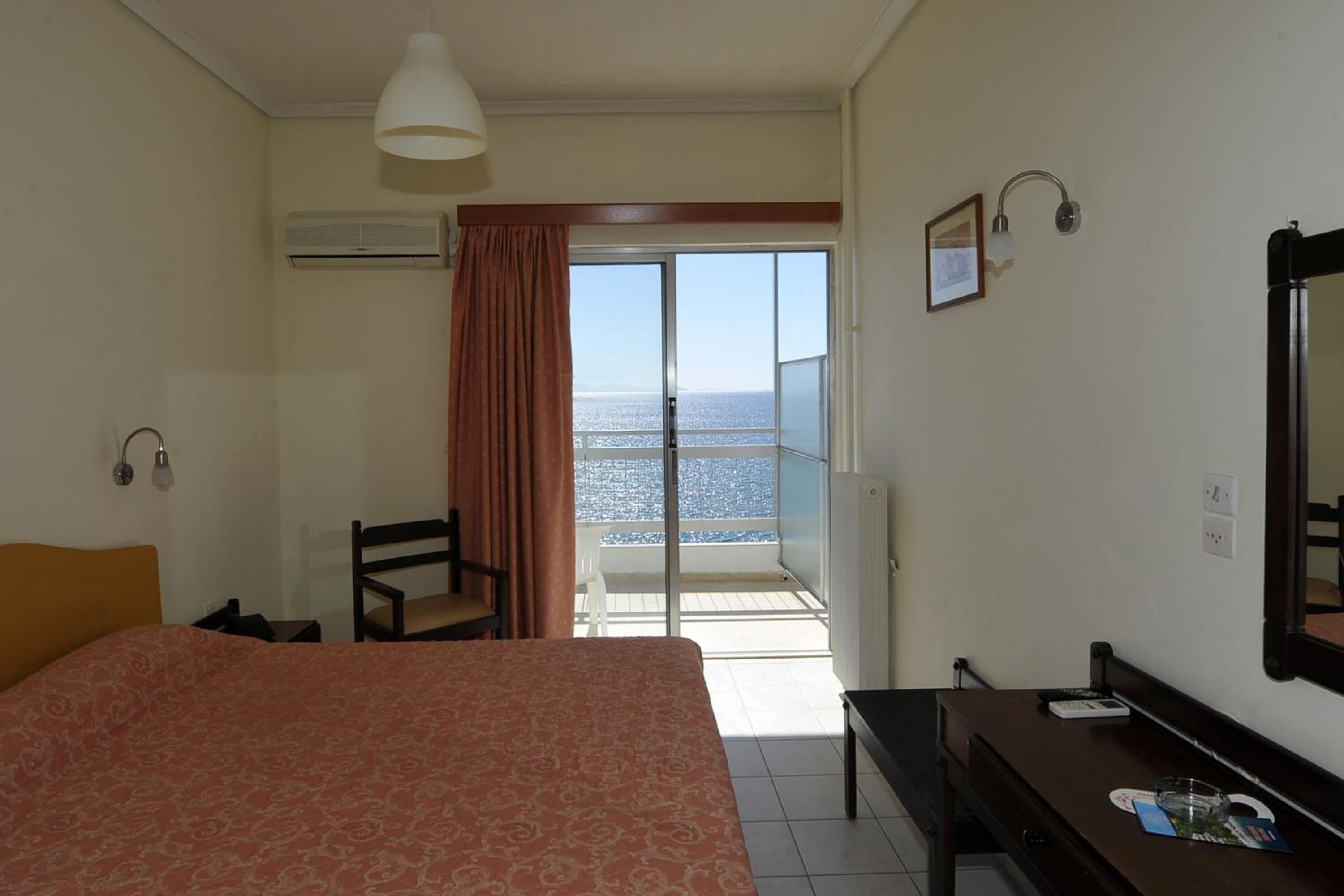 A hotel room with a bed, a desk, a chair, a large window with a view of the sea, and a balcony.