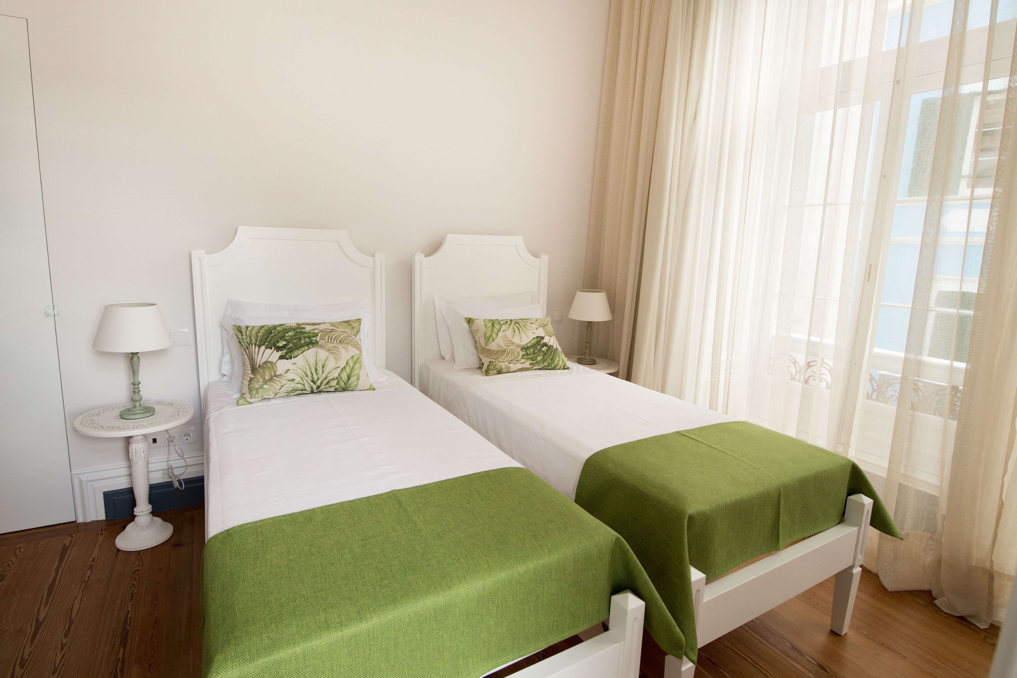 A hotel room with two single beds, a small round table with a lamp, and a window with sheer curtains.