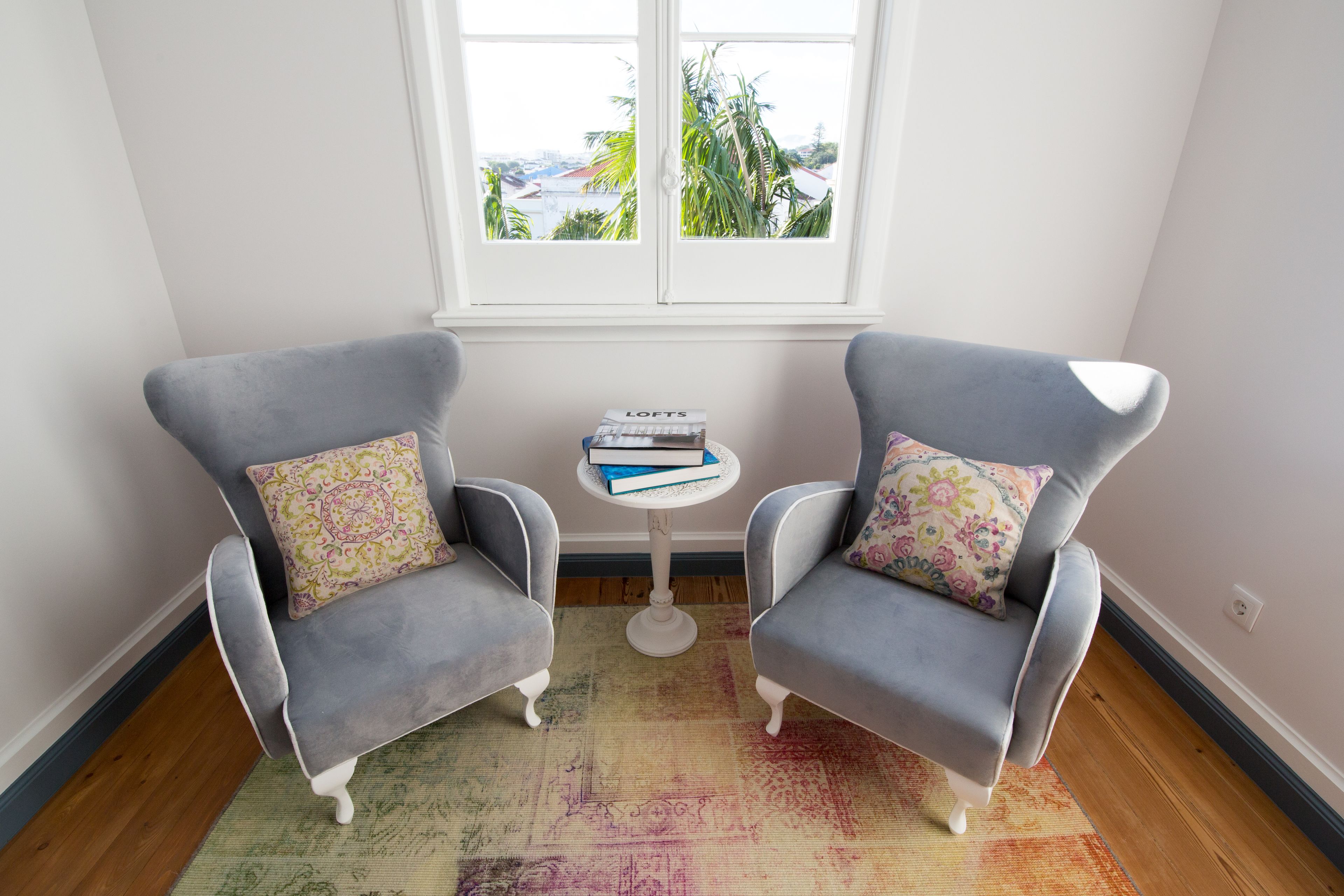 A corner room with two grey armchairs, a small white table with books, and a colorful rug.