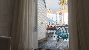 Una habitación con piso de madera, una puerta con cortinas blancas, y vista a un balcón con una silla azul y una palmera afuera.