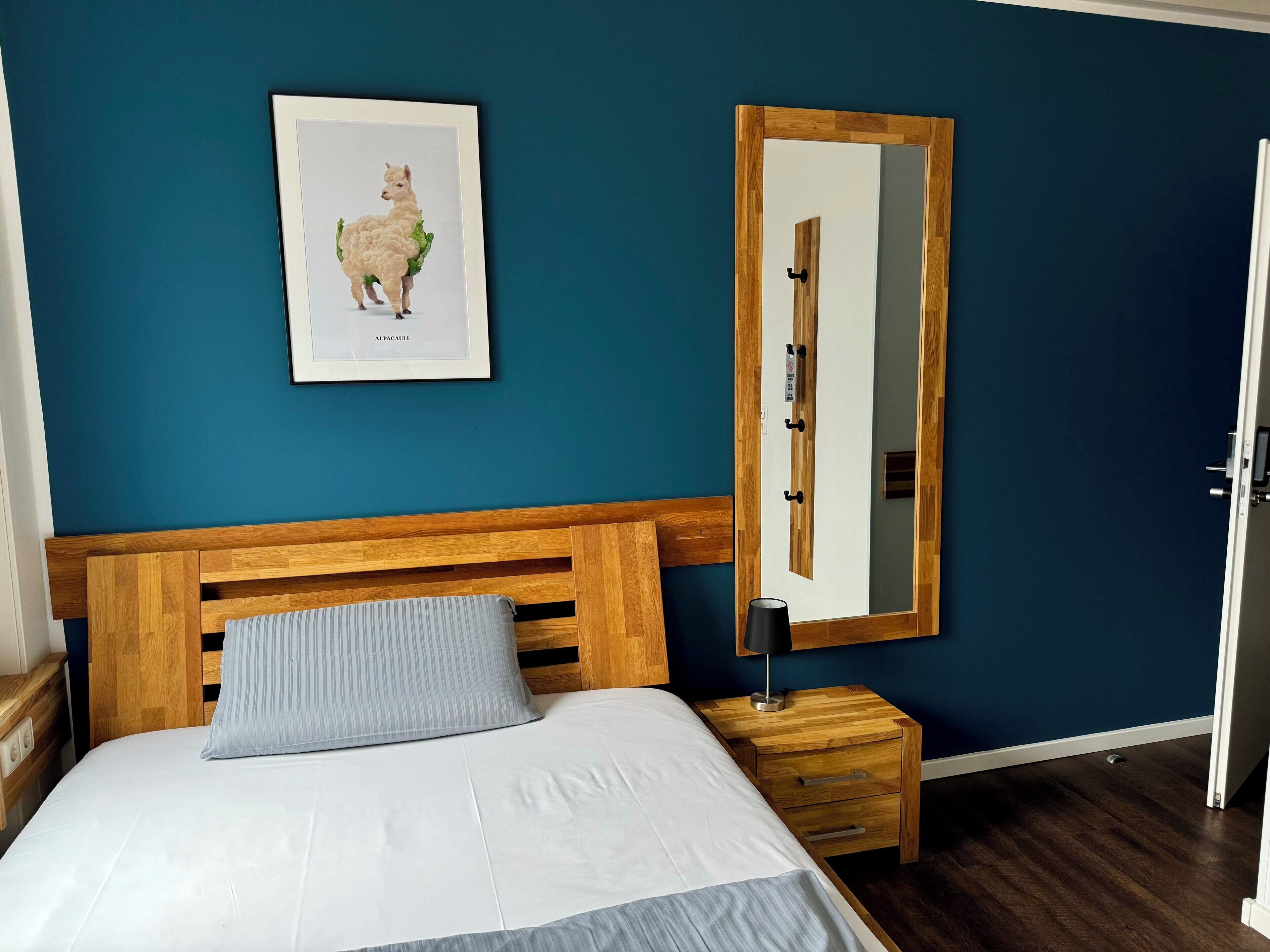 A bedroom with a wooden headboard, a bed with white bedding, a bedside table with a lamp, a framed picture of an alpaca, and a mirror on the wall.
