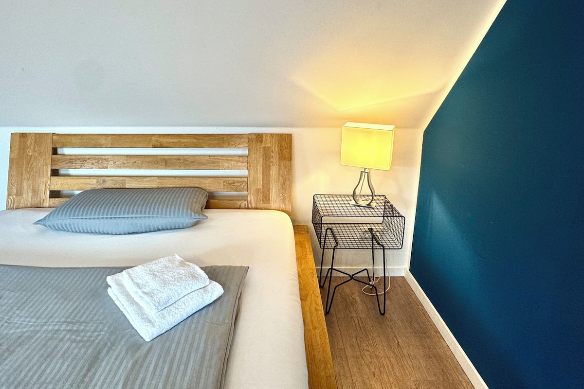 A neatly made bed with a wooden headboard, a single pillow, and a folded towel on the bed. There is a small bedside table with a lamp and a power outlet. The room has a blue accent wall and a wooden floor.