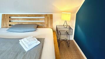 Un lit bien fait, avec une tête de lit en bois, un seul oreiller et une serviette pliée posée sur le lit. On y trouve une petite table de chevet avec une lampe et une prise électrique. La pièce est agrémentée d’un mur d’accent bleu et d’un plancher en bois.