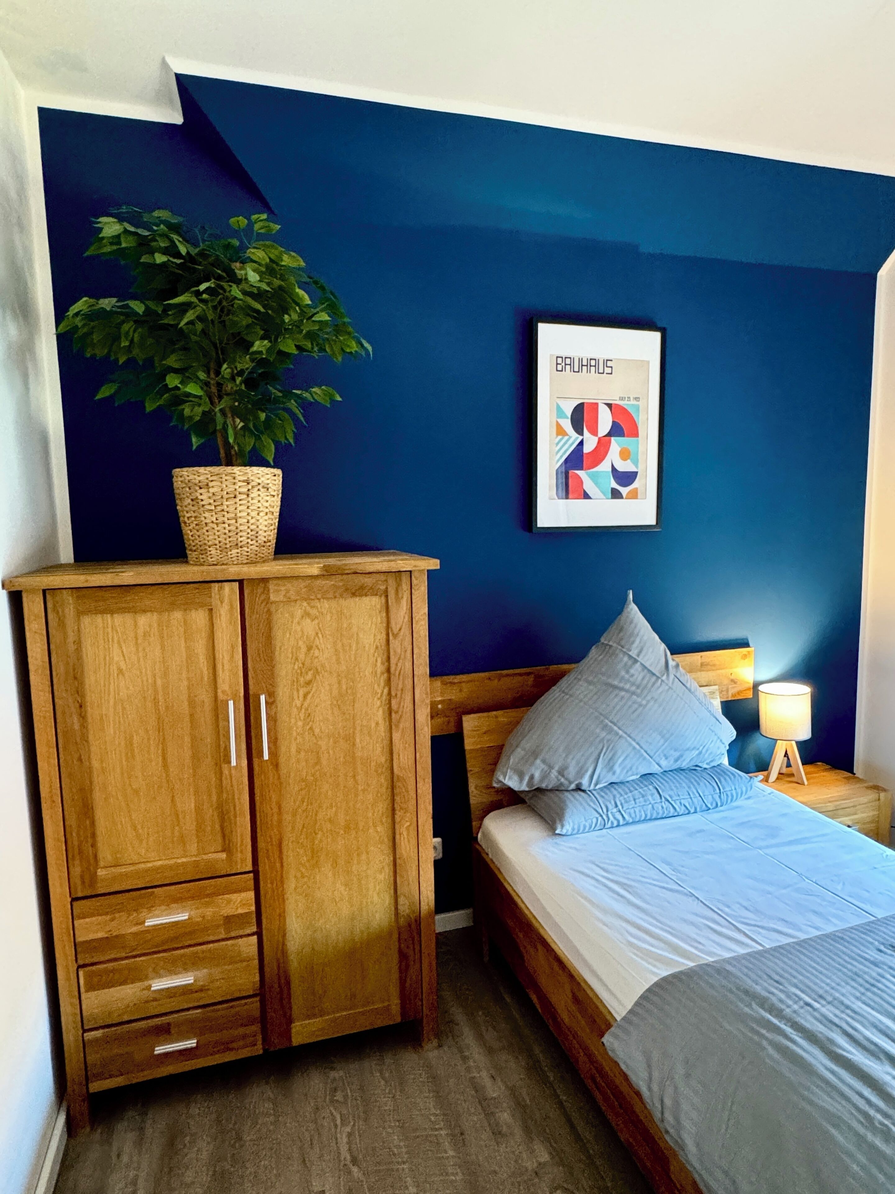 A bedroom with a wooden wardrobe, a bed with blue bedding, a nightstand with a lamp, and a framed abstract art piece on the wall.