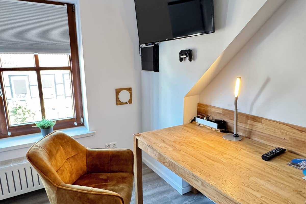 A modern room with a wooden desk, a comfortable chair, a flat-screen TV mounted on the wall, and a window with blinds.