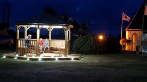 A gazebo with a wooden structure and benches, surrounded by grass and a small building with a flag. - Perran View Holiday Park (St. Agnes)