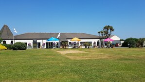 A building with a white facade, a grey roof, and a flagpole with a flag. There are colorful umbrellas and potted plants in front. - Perran View Holiday Park (St. Agnes)