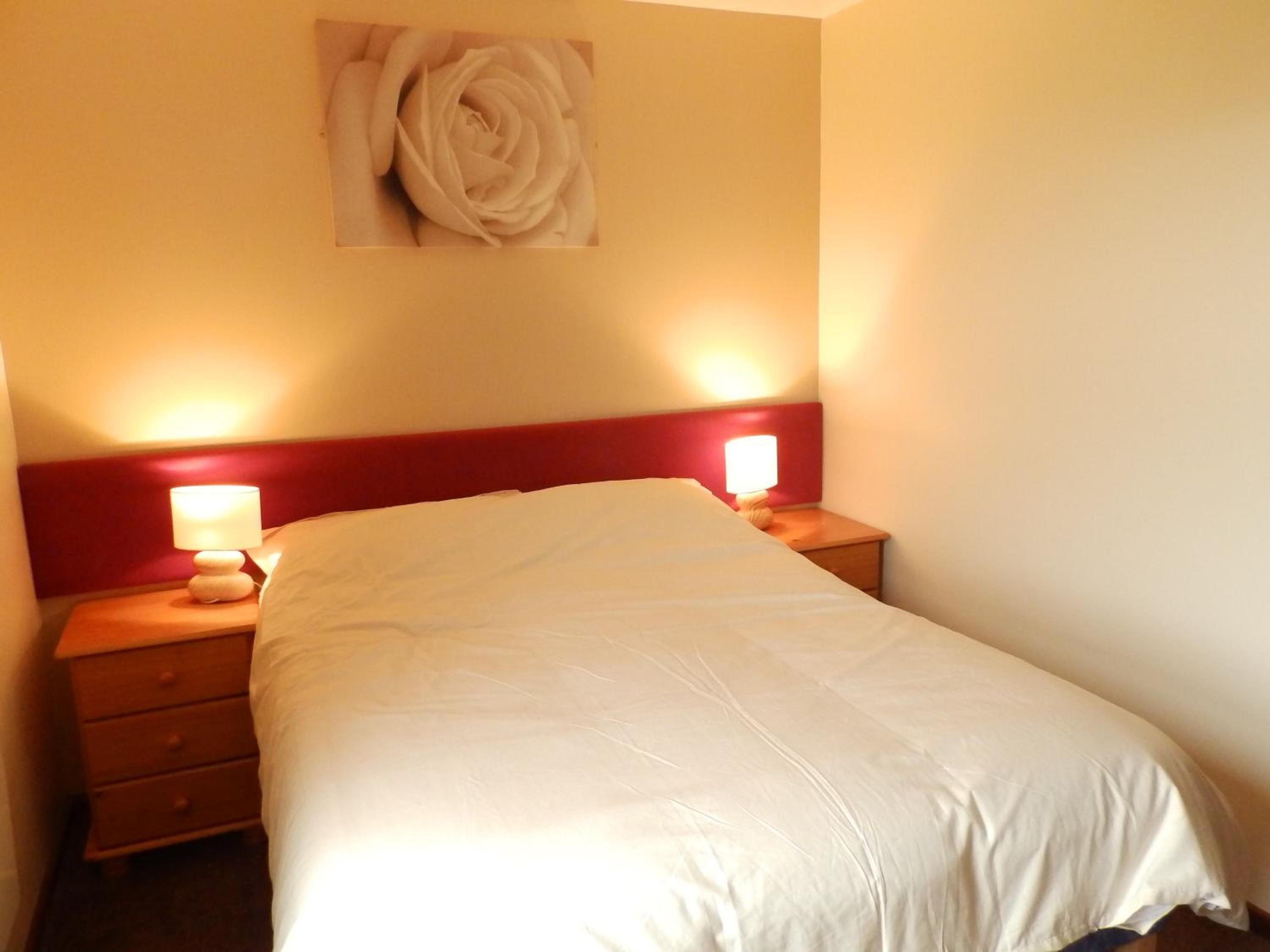 A neatly made bed in a hotel room with a red headboard, two bedside lamps, and a wall-mounted artwork of a rose.