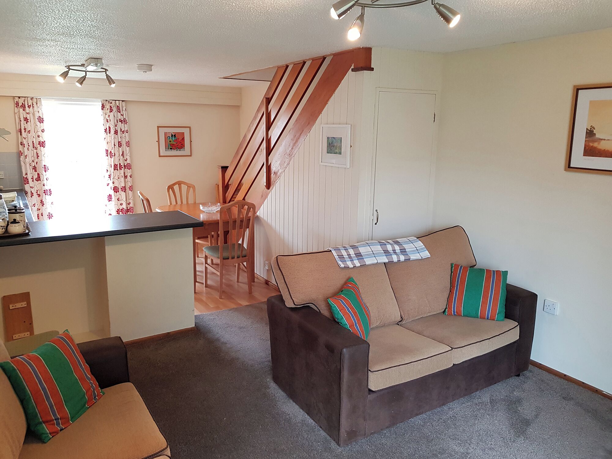 Una sala de estar con sofá, mesa de comedor, sillas, una escalera y una zona de cocina.