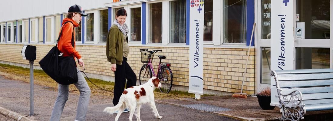 Una persona paseando un perro por una acera frente a un edificio con un letrero que dice "Välkommen!".