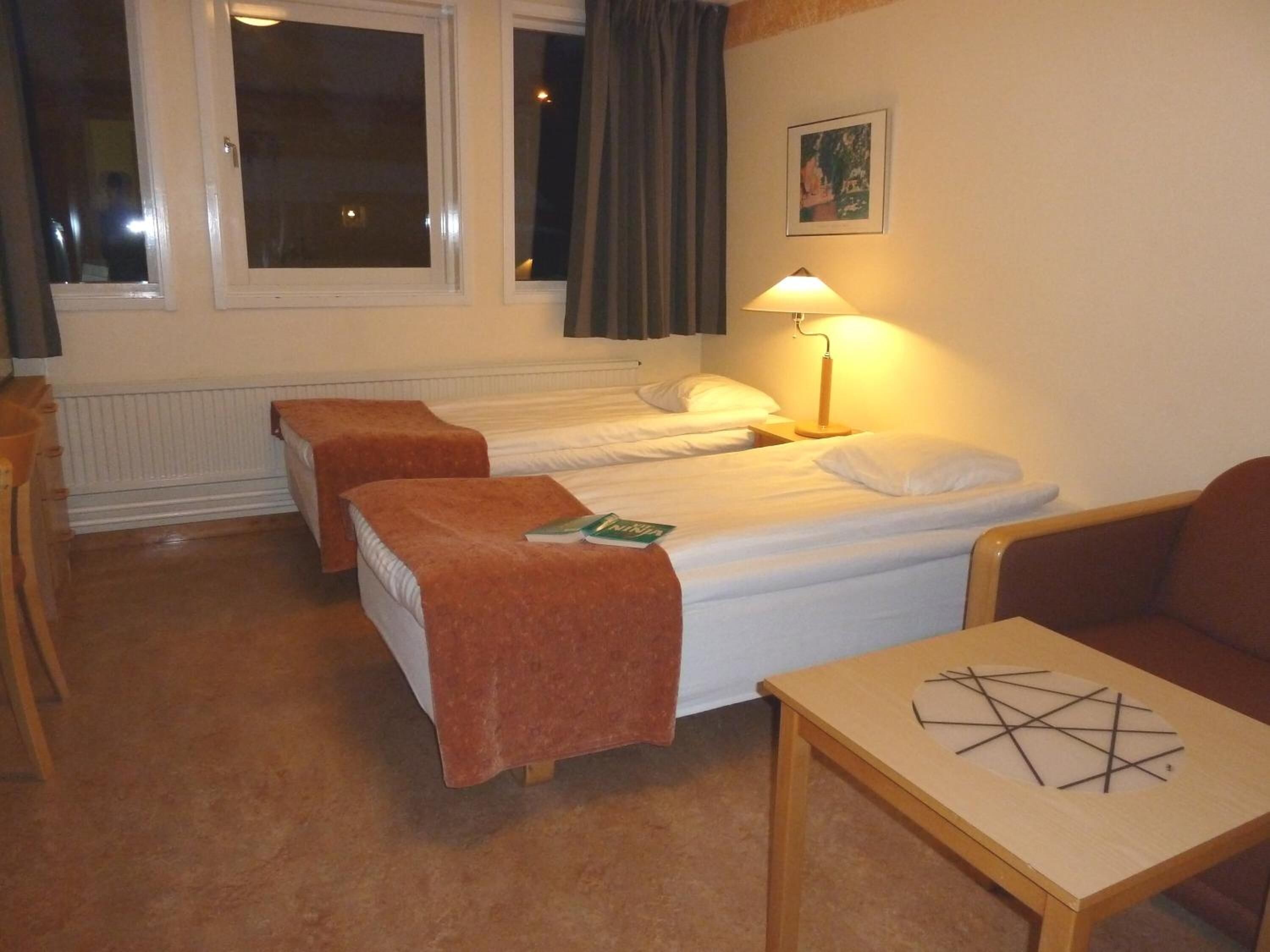 Habitación de hotel con dos camas individuales, una mesa de madera, una silla, una lámpara y un ventanal con cortinas.