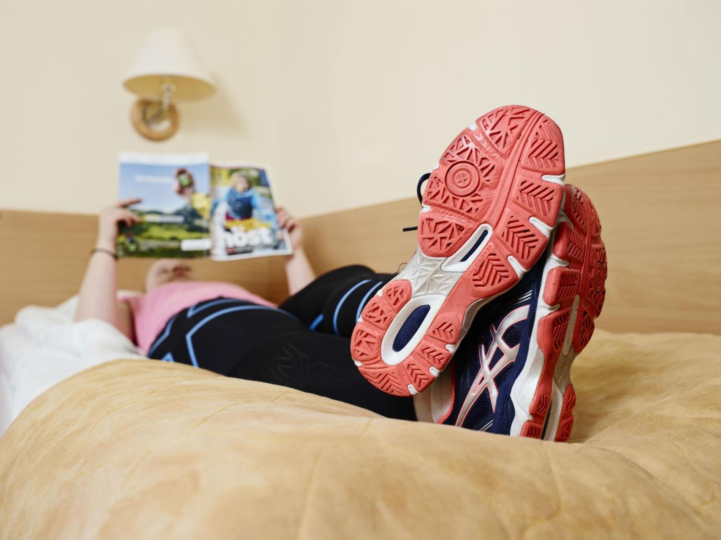 A person lying on a bed with a magazine, wearing a pair of athletic shoes with a distinctive red sole.