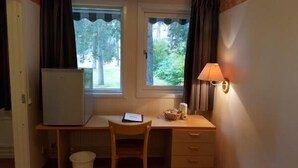 A small room with a desk, chair, and a lamp. There are two windows with curtains, a radiator, and a trash bin. - Hotell Sport & Rest (Pitea)