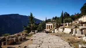 A historical site with ancient ruins, including stone steps leading up to a temple-like structure, surrounded by trees and mountains.