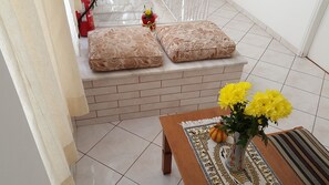 A room with a tiled floor, a wooden bench, two patterned cushions on a bench, a vase with yellow flowers, and a red fire extinguisher.