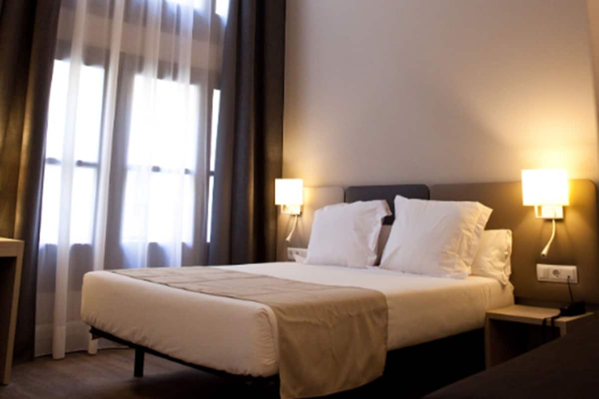 A hotel room with a bed, two bedside lamps, a nightstand, and a window with curtains.