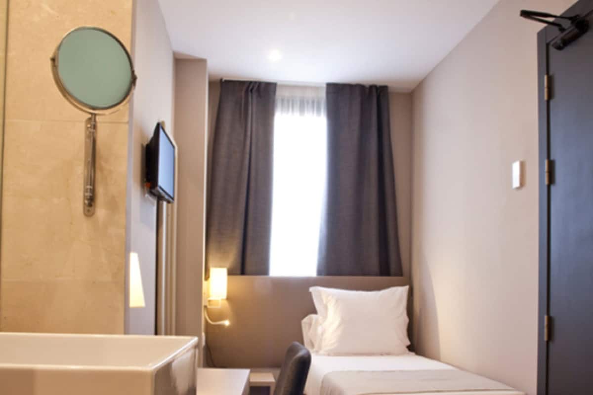A compact hotel room with a bed, a desk, a chair, a small bathroom with a mirror, and a TV.