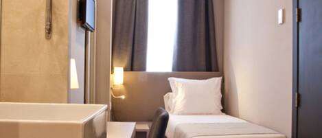 A compact hotel room with a bed, a desk, a chair, a small bathroom with a mirror, and a TV.