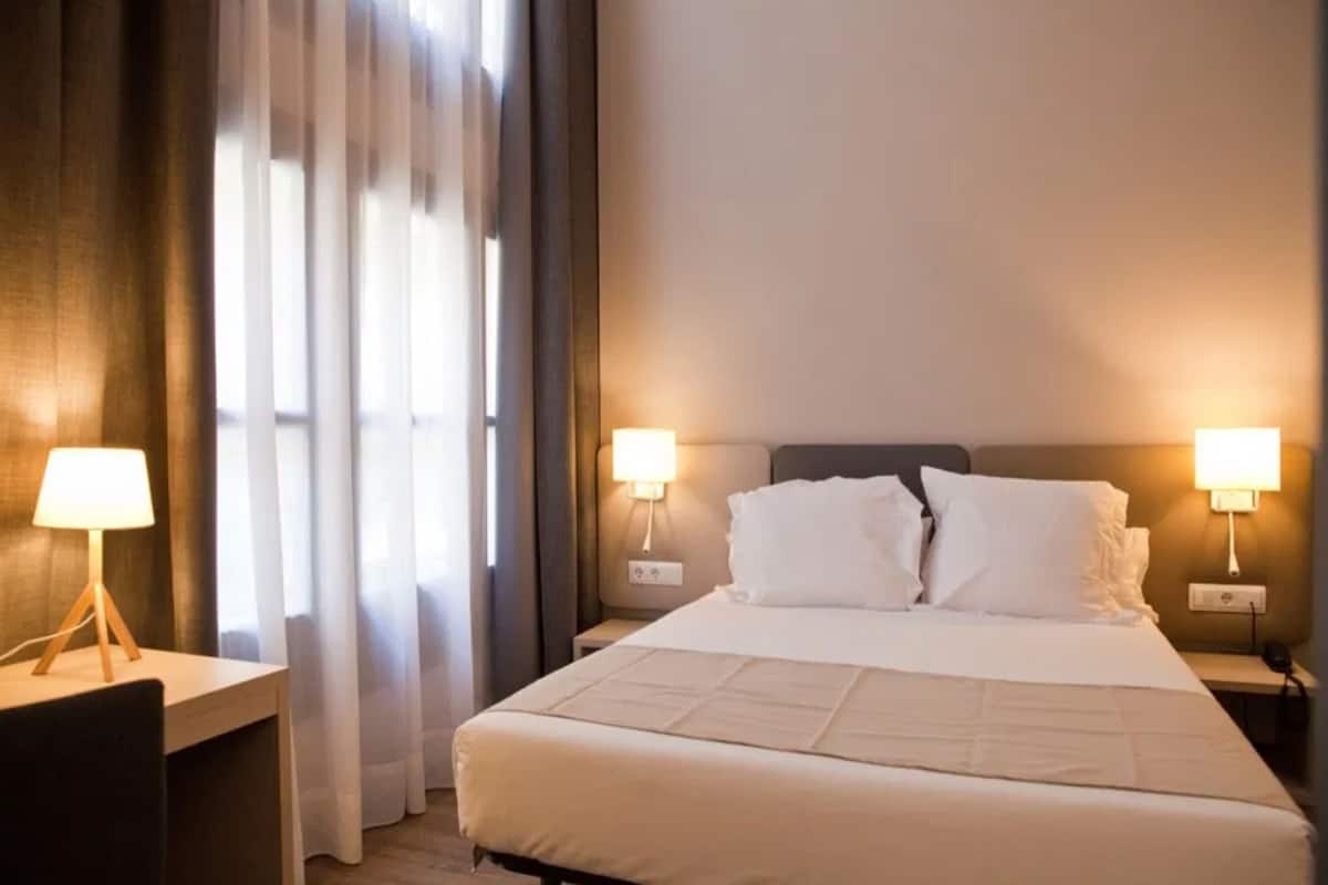 A hotel room with a bed, two bedside lamps, a desk with a lamp, and a window with curtains.