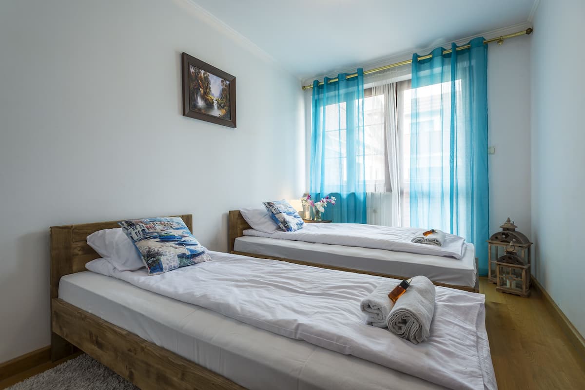 A room with two single beds, a wooden headboard, a framed picture on the wall, a window with blue curtains, and a small lantern on the floor.