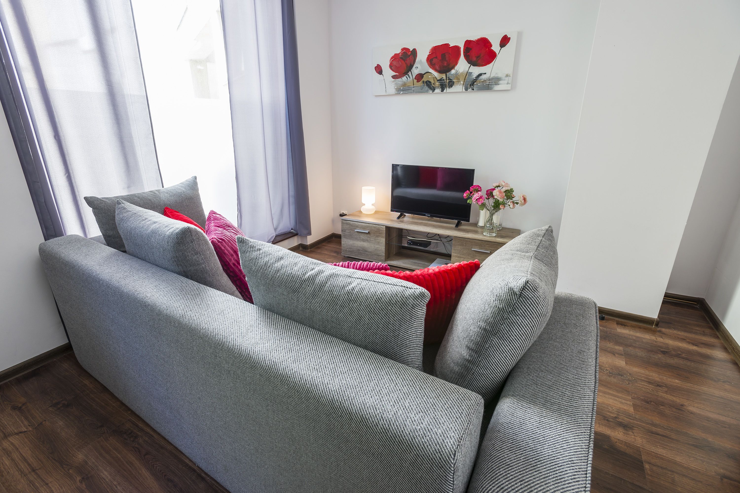 A modern living room with a grey sofa, a flat-screen TV, and a small table with a vase of flowers.