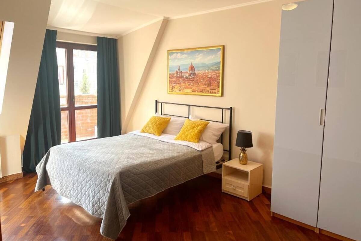 A bedroom with a bed, a nightstand, a lamp, a wardrobe, a painting, and a window with curtains.