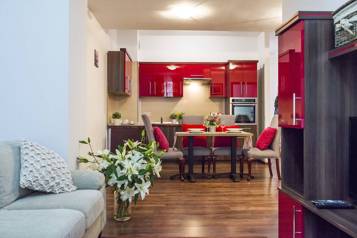 A modern living room with a grey sofa, a dining table with chairs, and a kitchen in the background with red cabinets.