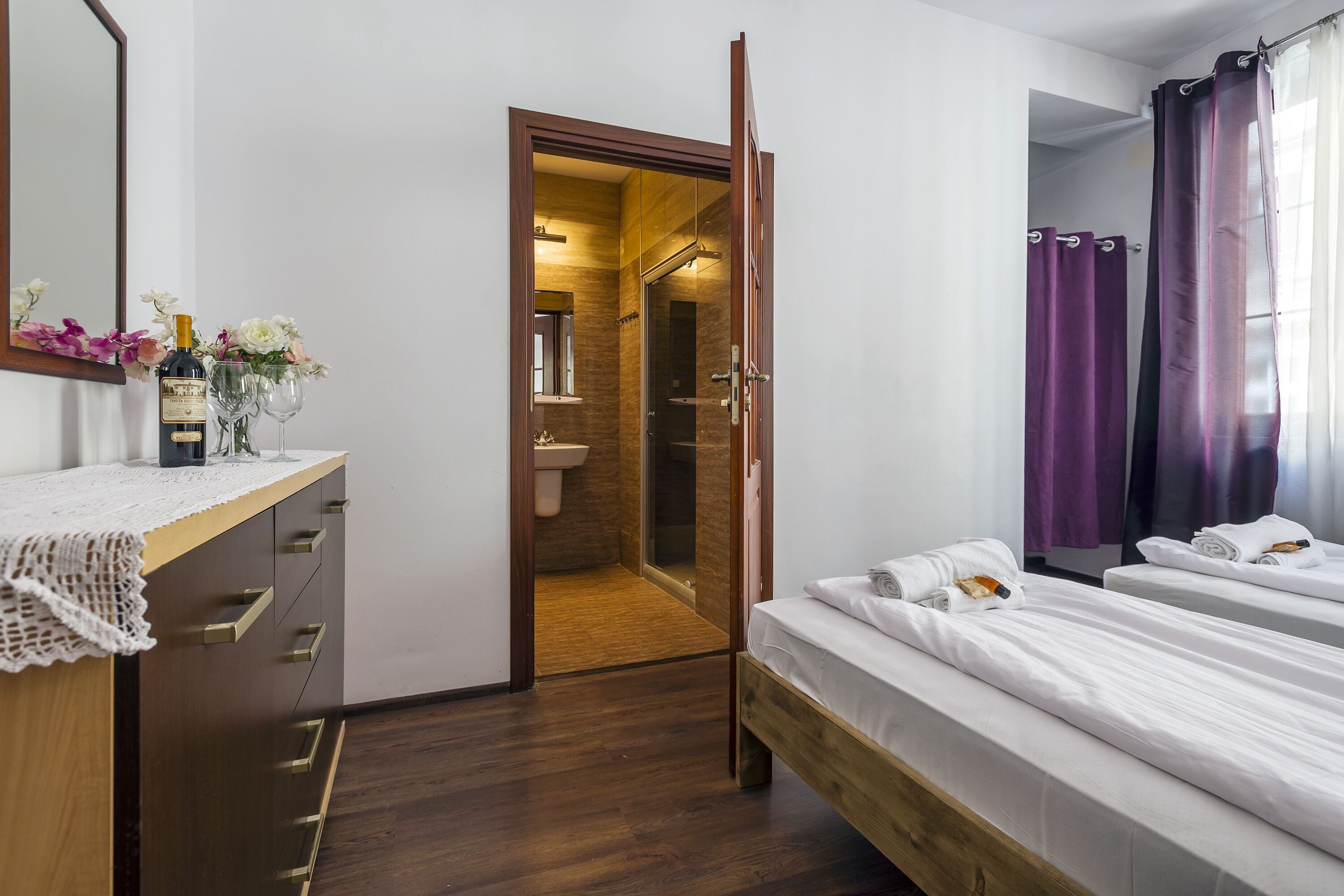 A hotel room with a wooden bed, a bedside table with a bottle and glasses, a mirror, and a bathroom visible through an open door.