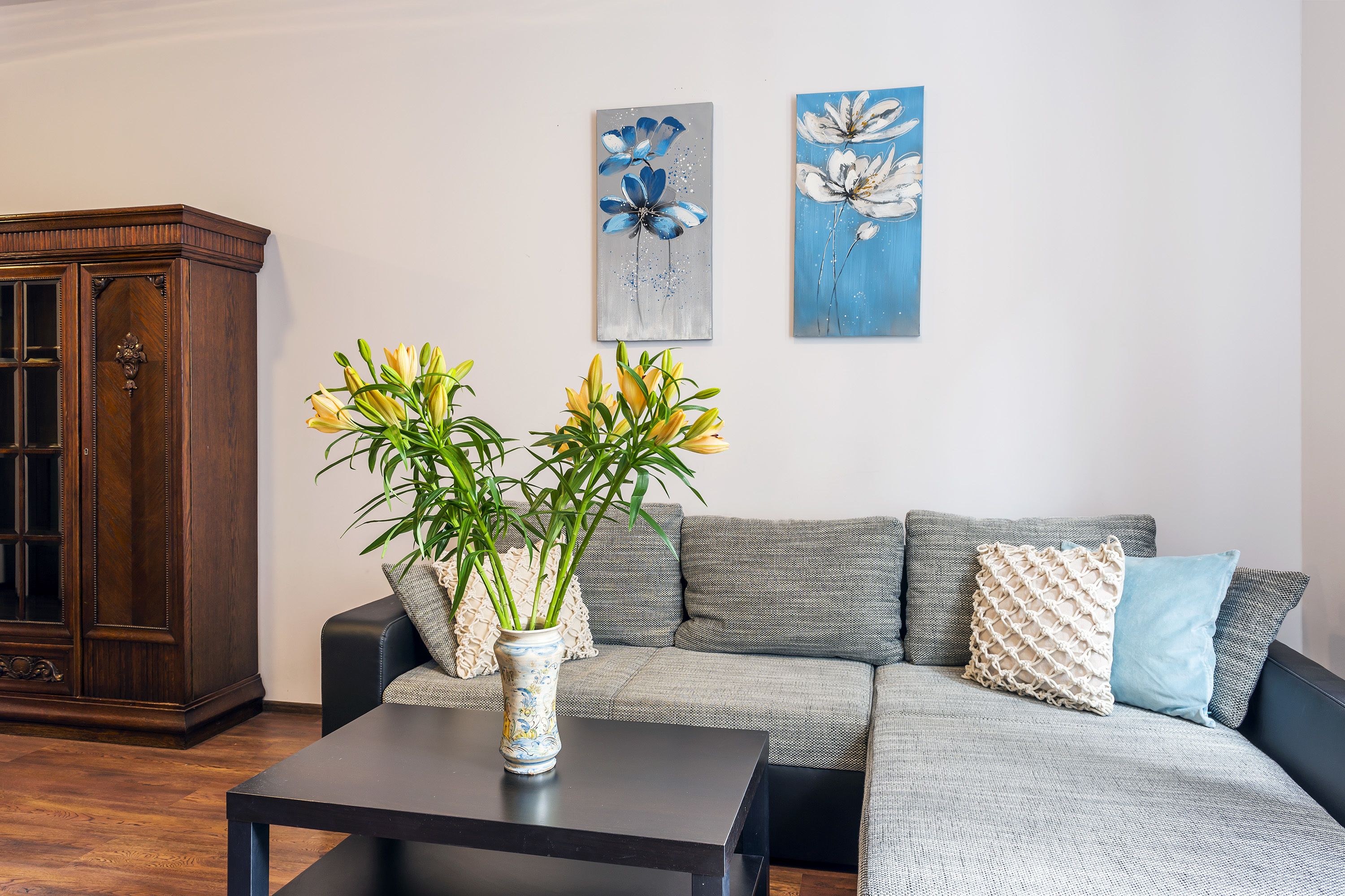 Un salón con un mueble de madera oscura, un sofá gris con cojines estampados y un jarrón con flores amarillas sobre una mesa negra.