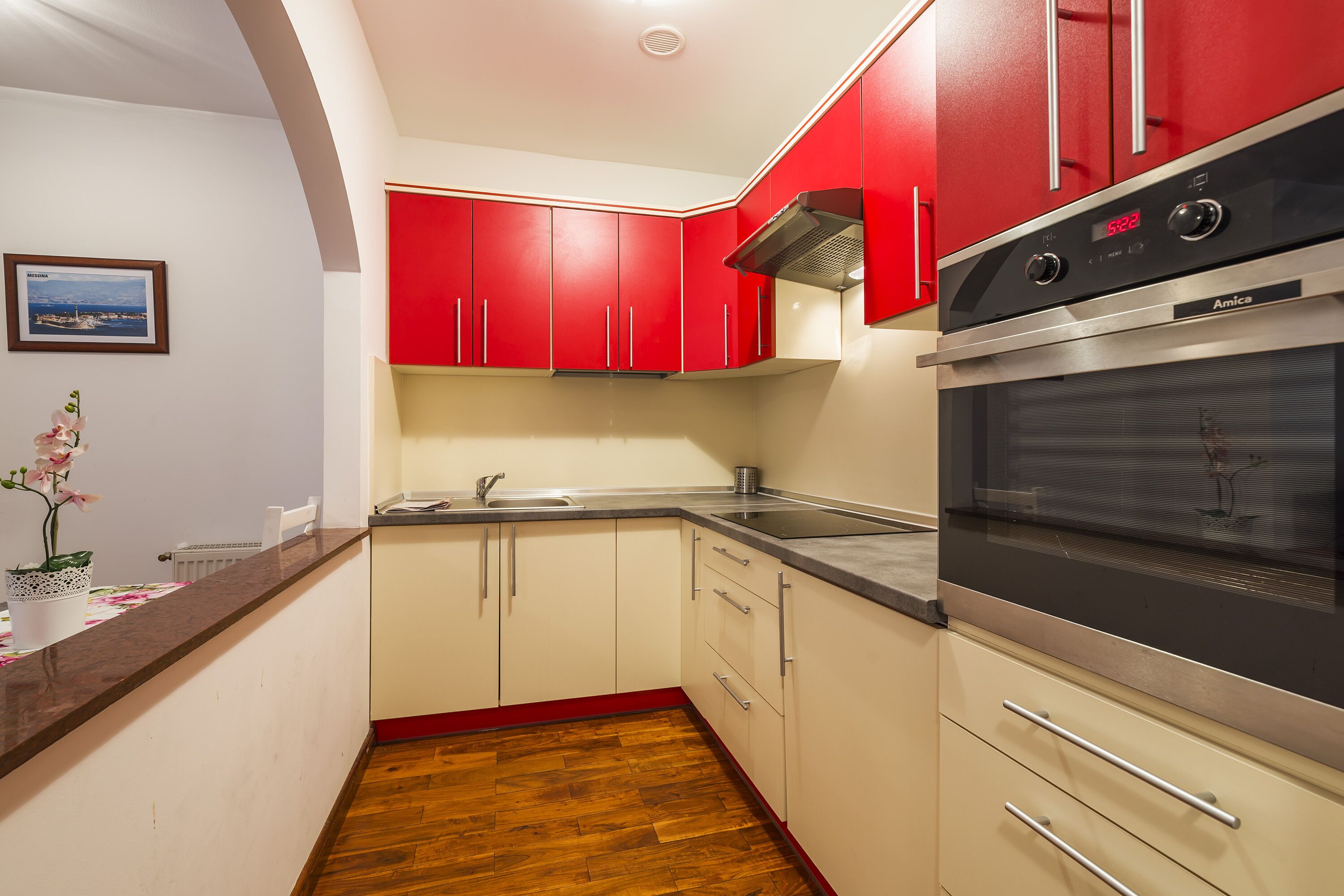 A kitchen with red cabinets, a stainless steel oven, and a wooden floor.