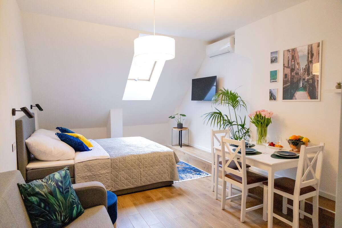 A compact bedroom with a bed, a small seating area, a dining table with chairs, and a large window providing natural light.
