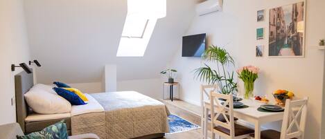 A compact bedroom with a bed, a small seating area, a dining table with chairs, and a large window providing natural light.