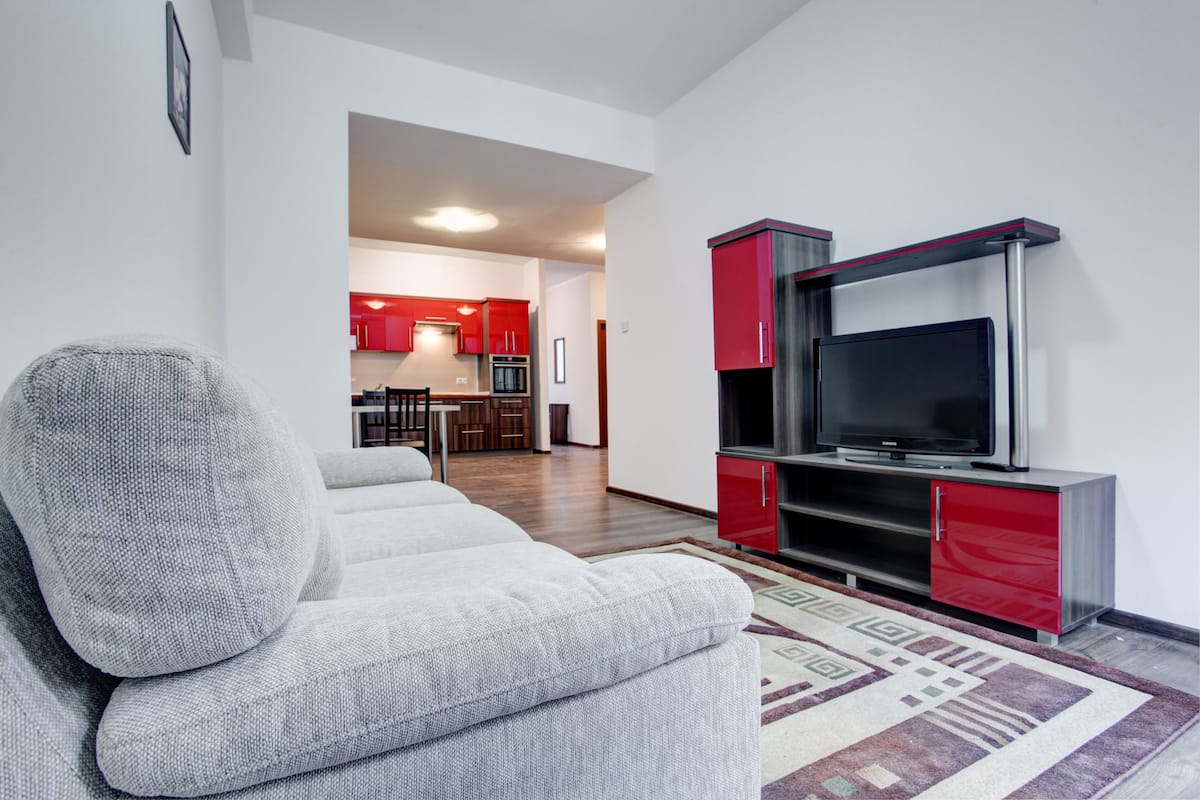 A modern living room with a grey sofa, a flat-screen TV on a red and black entertainment unit, and a patterned rug.